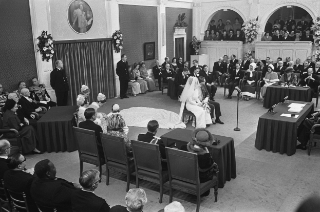 Ein Schwarz-Weiß-Foto einer Hochzeitszeremonie in einem großen Raum mit Gästen auf Stühlen und Tischen mit Papieren und Mikrofonen, einer dekorierten Wand mit Fotorahmen und Blumenbouquets und einem Podium mit Mikrofonständer.