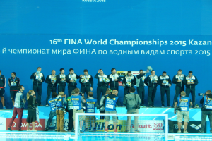 Gruppe von Menschen auf einem Schwimmbaddeck mit Kameras, einem Netz im Vordergrund und einem "FINA World Championships 2015 Kazan"-Schild im Hintergrund.