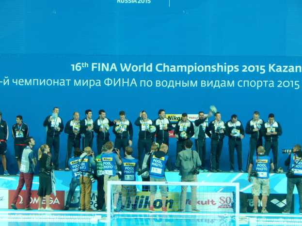 Gruppe von Menschen auf einem Schwimmbaddeck mit Kameras, einem Netz im Vordergrund und einem "FINA World Championships 2015 Kazan"-Schild im Hintergrund.
