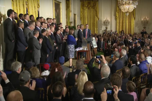 Eine Gruppe von Menschen mit Kameras steht vor einer Menge, einem Podium mit Mikrofon, einer Tafel mit Text, einem Topf mit Pflanze, Kerzen, Rahmen, Vorhängen und einem Kronleuchter, vermutlich fotografieren sie den Präsidenten und Vizepräsidenten der USA.