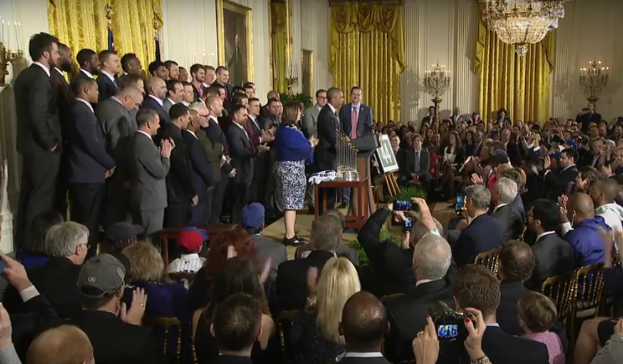 Eine Gruppe von Menschen mit Kameras steht vor einer Menge, einem Podium mit Mikrofon, einer Tafel mit Text, einem Topf mit Pflanze, Kerzen, Rahmen, Vorhängen und einem Kronleuchter, vermutlich fotografieren sie den Präsidenten und Vizepräsidenten der USA.
