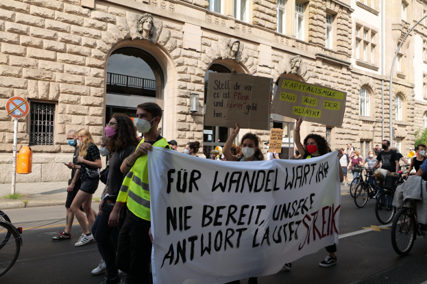 Eine Gruppe von Menschen marschiert bei einer Protestaktion gegen die Anti-Kriegsbewegung in Berlin, einige halten Schilder und andere fahren Fahrräder, im Hintergrund ein Gebäude mit Fenstern, Bögen, Säulen und Skulpturen.