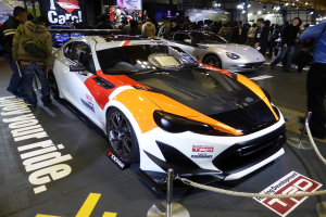 Eine Gruppe von Menschen um ein Rennfahrzeug auf der Tokyo Motor Show herumsteht, umgeben von Absperrpollern mit Seilen, mit Informationsschildern im Hintergrund unter beleuchteter Decke.