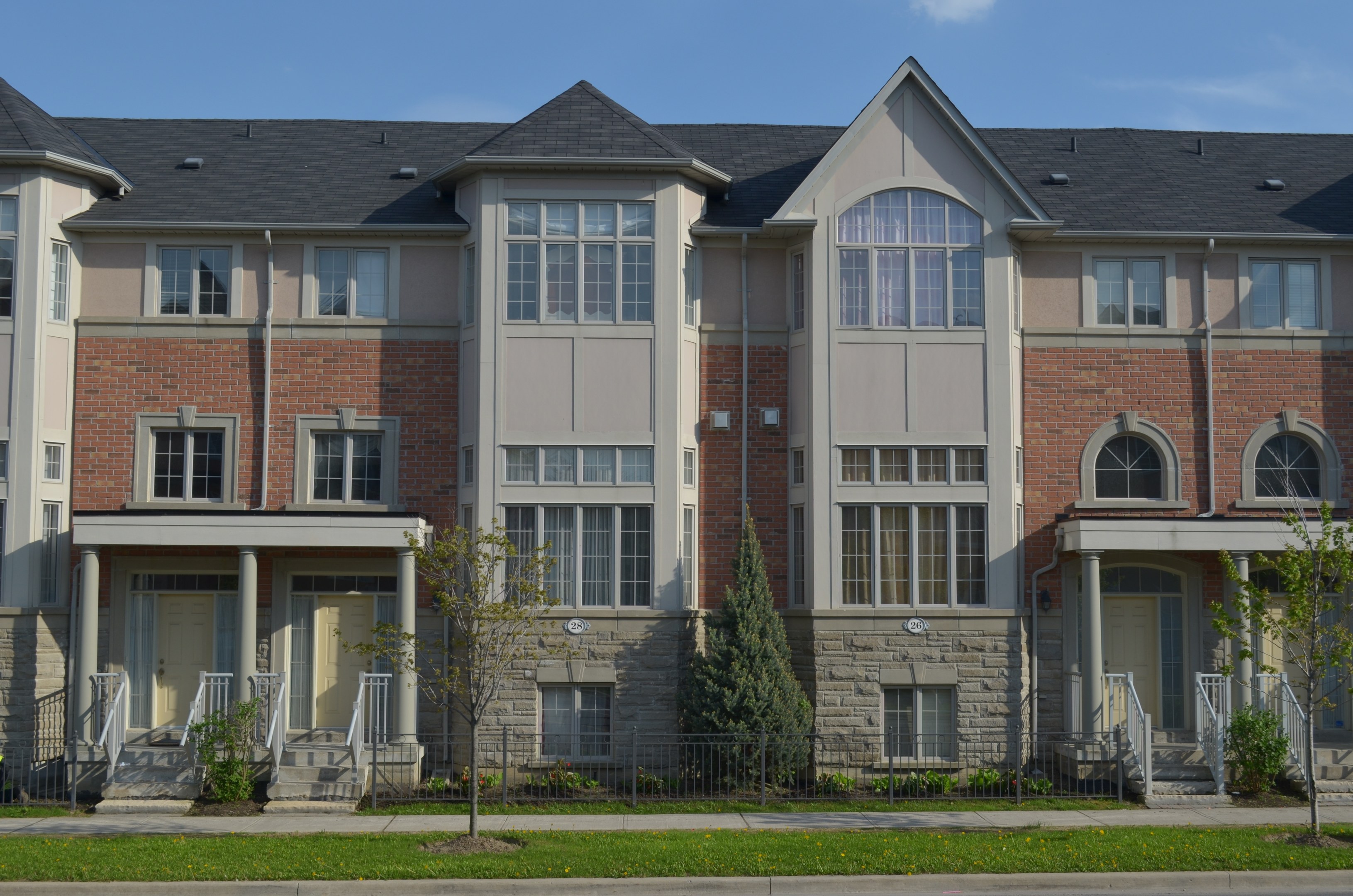 Eine Reihe von Stadthäusern in einem Wohnviertel mit Fenstern, Türen, Säulen, Geländern, Treppen, Pflanzen, Bäumen, Gras, einer Straße im Vordergrund und dem Himmel im Hintergrund.