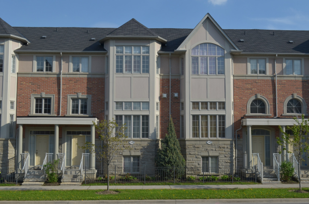 Eine Reihe von Stadthäusern in einem Wohnviertel mit Fenstern, Türen, Säulen, Geländern, Treppen, Pflanzen, Bäumen, Gras, einer Straße im Vordergrund und dem Himmel im Hintergrund.