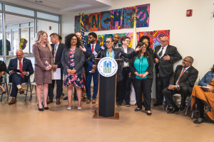 Eine Gruppe von Menschen steht vor einem Rednerpult mit einem Mikrofon und einer Tafel mit der Aufschrift "Sancho-Stadtrat tritt dem Stadtrat bei", wobei einige sitzen und andere stehen, und ein Hintergrund mit Flaggen, einer Tafel, einem Feuerlöscher und Deckenleuchten.