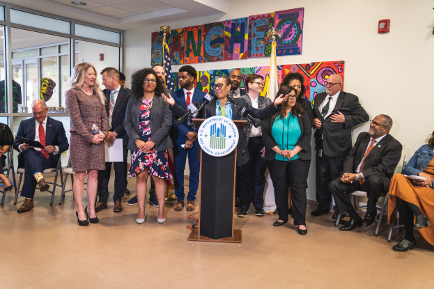 Eine Gruppe von Menschen steht vor einem Rednerpult mit einem Mikrofon und einer Tafel mit der Aufschrift "Sancho-Stadtrat tritt dem Stadtrat bei", wobei einige sitzen und andere stehen, und ein Hintergrund mit Flaggen, einer Tafel, einem Feuerlöscher und Deckenleuchten.