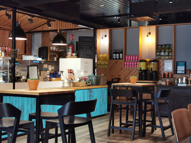 Inneneinrichtung eines Restaurants mit Holztischen, Stühlen, einem Topf mit einer Pflanze auf einem Tisch, einer Schiefertafel, einem Kühlschrank, einem Feuerlöscher, Regalen mit Flaschen, Schränken mit Gegenständen, einem Rahmen mit einem Foto an der Wand und Deckenleuchten.