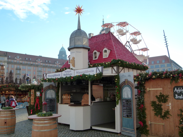 Ein kleines Gebäude mit rotem Dach und einem Riesenrad davor, umgeben von Festdekorationen und Menschen, mit Gebäuden, Bäumen und Wolken im Hintergrund auf einem Oktoberfest-Event in München, Deutschland.