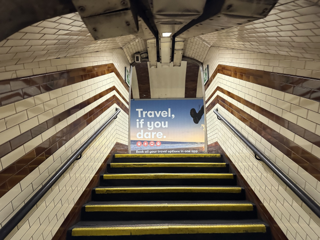 Eine Treppe in einer U-Bahn-Station mit einem 'Reise, wenn du wagst'-Schild oben, flankiert von Geländern und sichtbaren Stationswänden im Hintergrund.