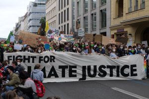 Eine große Gruppe von Menschen marschiert eine Straße entlang, hält ein Banner mit der Aufschrift "Kämpfen für Hassjustiz" in den Händen, wobei einige Masken tragen und Schilder oder Taschen tragen, vor einem Hintergrund von Gebäuden, Bäumen und einem klaren blauen Himmel.