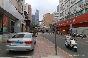 Ein Mann fährt mit einem Roller eine Straße mit hohen Gebäuden entlang, mit Menschen auf den Bürgersteigen und Fahrzeugen auf der Straße, unter einem klaren blauen Himmel mit Bäumen im Hintergrund.