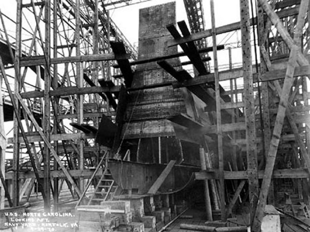 Schwarzes und weißes Foto eines Schiffs in einem Schiffbauwerk mit Holzpfählen und Treppen im Vordergrund und Himmel im Hintergrund, mit dem Text "Marinebewaffnung" in der linken unteren Ecke.
