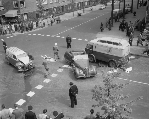 Eine Schwarz-Weiß-Szene eines Autounfalls an einer Straßecke mit mehreren Fahrzeugen, darunter ein Lieferwagen, einer Gruppe von Menschen drumherum und im Hintergrund Gebäude, Bäume, Masten und Texttafeln.