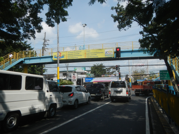 Vielbefahrene Straße mit Fahrzeugen, eine Brücke mit Geländern und Treppen, Laternen, Verkehrszeichen, Texttafeln, Bäume, Gebäude und ein bewölkter Himmel.
