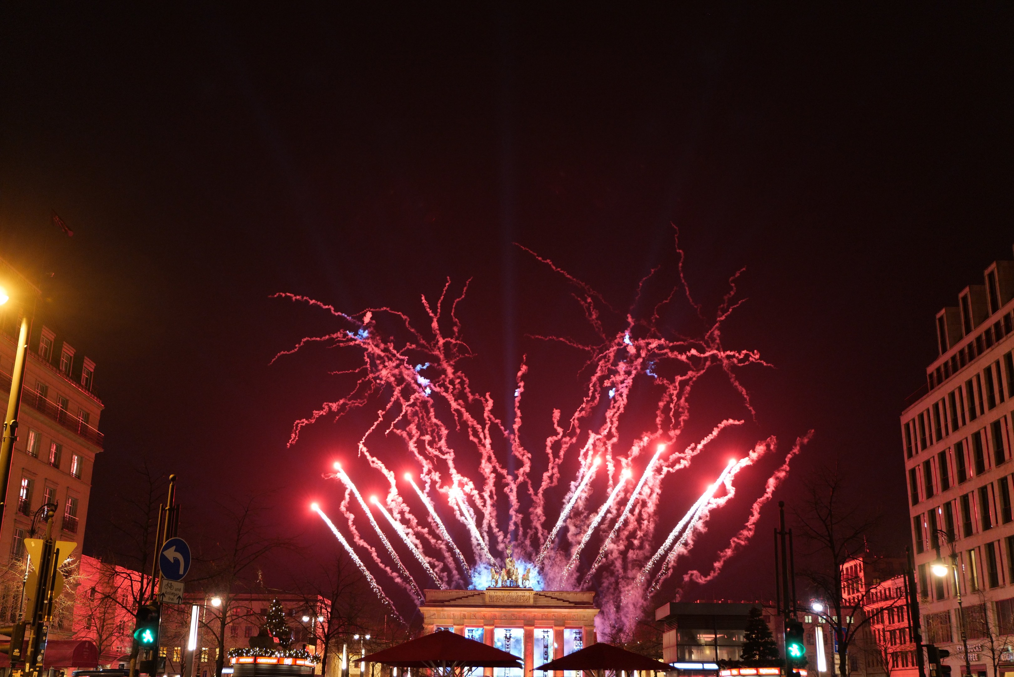 Eine belebte Stadtstraße an Silvester in Berlin, voller Menschen, Fahrzeuge und festliche Lichter von Gebäuden und Feuerwerk am Himmel.