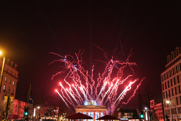 Eine belebte Stadtstraße an Silvester in Berlin, voller Menschen, Fahrzeuge und festliche Lichter von Gebäuden und Feuerwerk am Himmel.