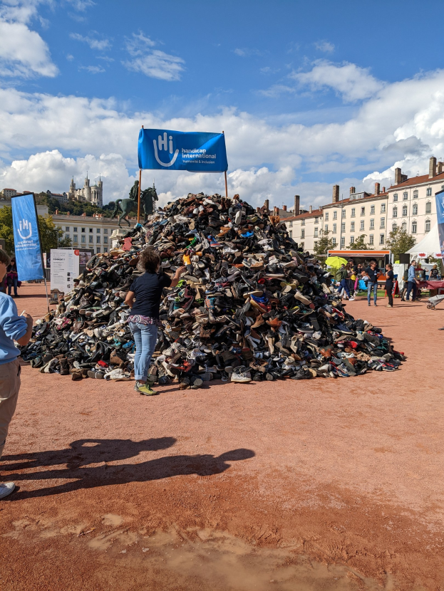 Eine Gruppe von Menschen steht um einen Haufen Schuhe auf einem Schmutzfeld mit Gebäuden, Bäumen und einem bewölkten Himmel im Hintergrund bei einem internationalen Schuh-Recycling-Event.