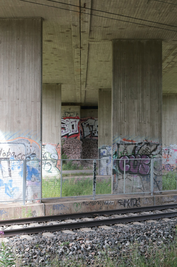Eisenbahnschiene mit Graffiti, umgeben von Steinen, Gras, einem Zaun, Säulen, Drähten und einem Dach.